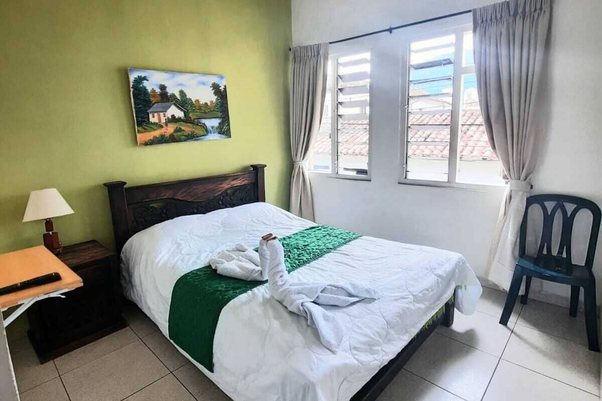 Ein Hotelzimmer mit Bett, Nachttisch, Stuhl, einem Fenster mit Vorhängen und einem Bild an der Wand.