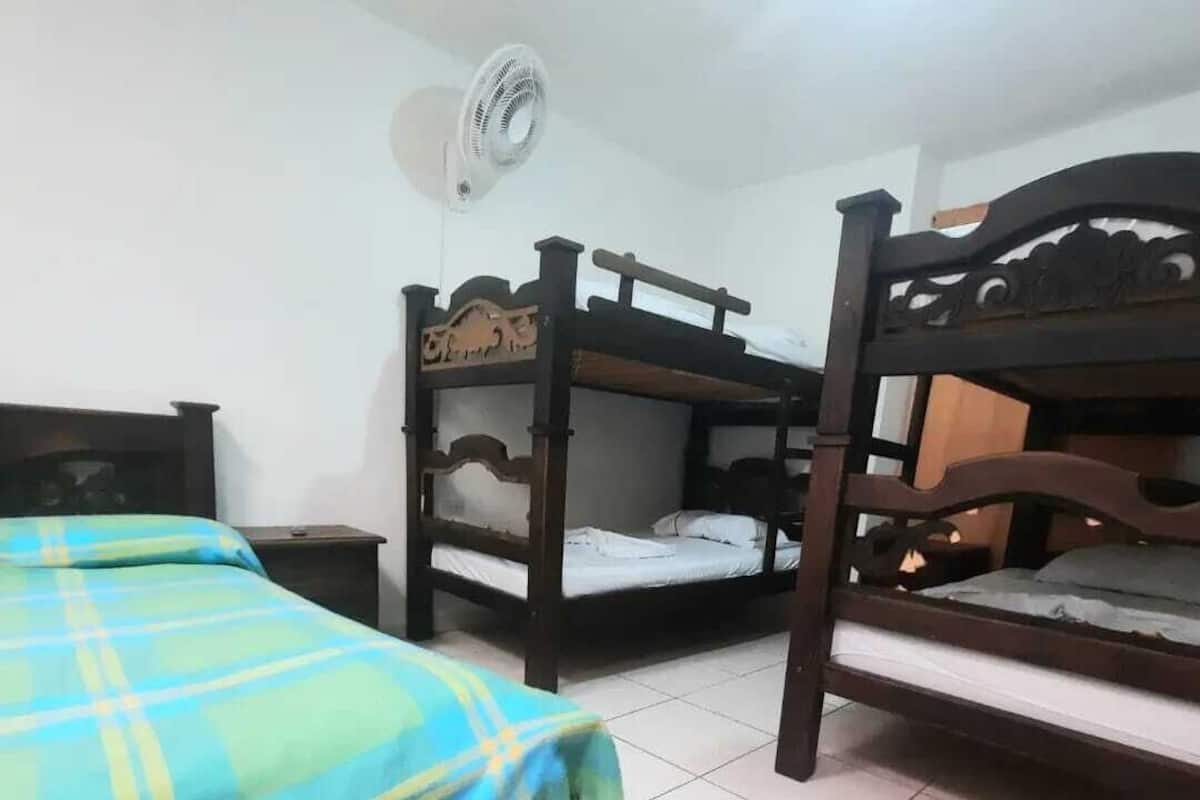 Ein Zimmer mit zwei Etagenbetten, einem Einzelbett mit Karo-Decke, einem kleinen Nachttisch und einem Deckenventilator.