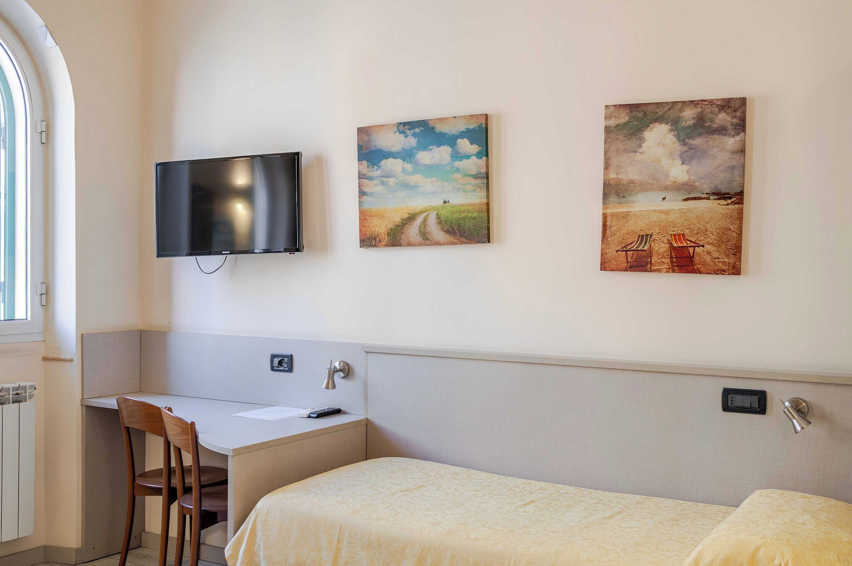 Hotellihuone, jossa on työpöytä, tuoli, sänky ja kaksi taulua seinällä.
