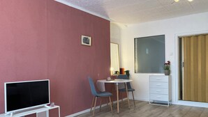 Habitación con televisor de pantalla plana sobre un soporte blanco, una mesita con dos sillas, un espejo y un armario con una planta.