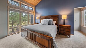 A bedroom with a large bed, wooden headboard, two bedside tables with lamps, and large windows offering a view of trees. - TITL08 - Fantastic Sunriver Home, Hot Tub (Sunriver)
