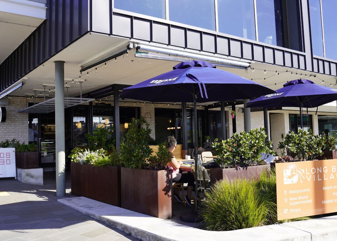 An outdoor seating area with umbrellas, a woman sitting, and a signboard listing amenities like restaurants and a supermarket.