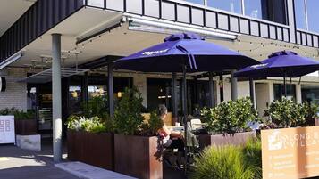 An outdoor seating area with umbrellas, a woman sitting, and a signboard listing amenities like restaurants and a supermarket.