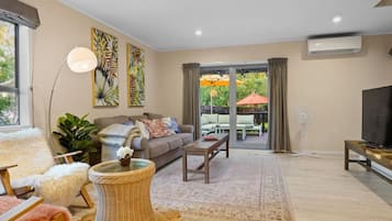 A modern living room with a sofa, coffee table, and TV. There is a large window and a sliding glass door leading to an outdoor patio.
