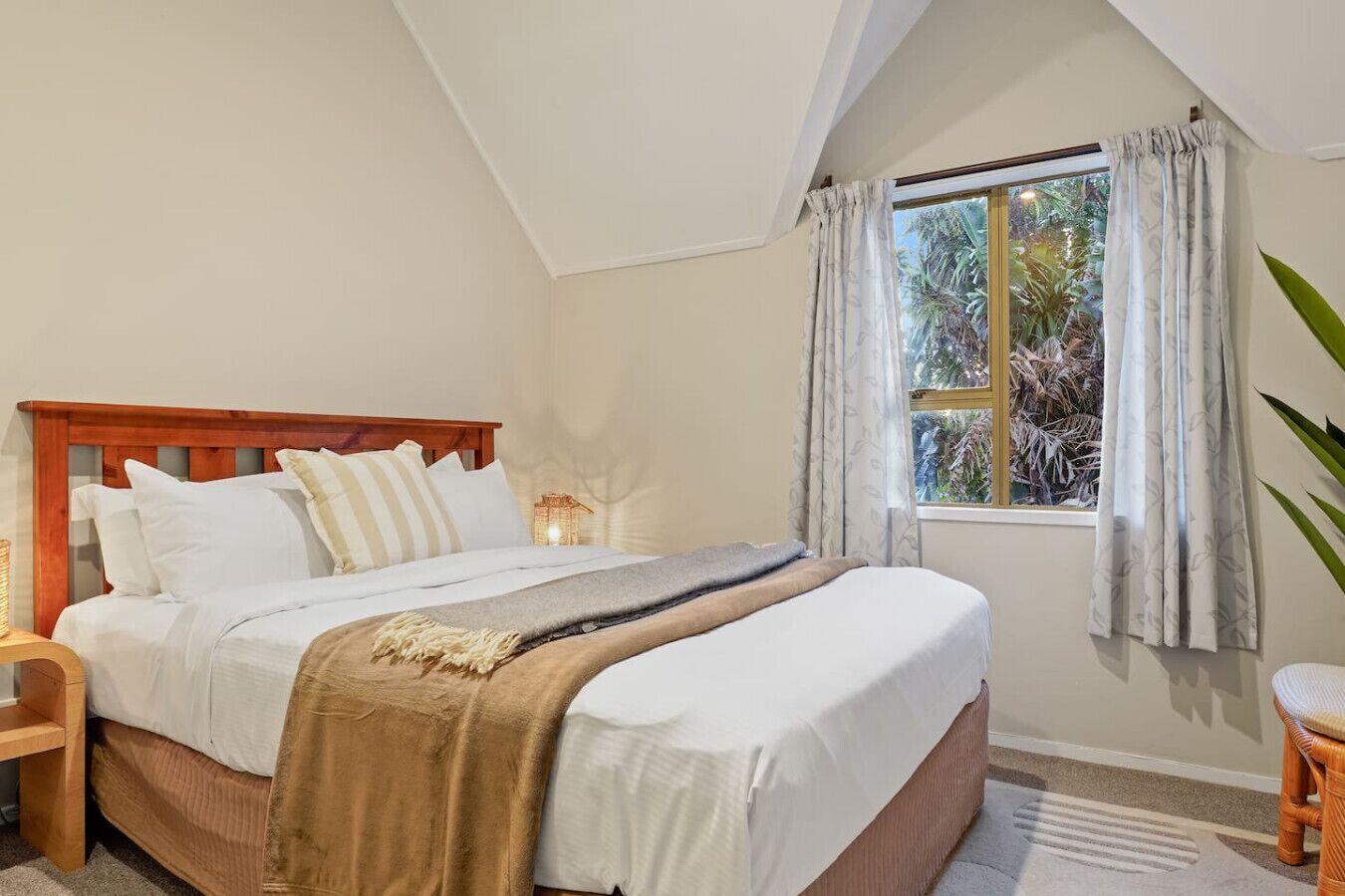 A bedroom with a wooden bed frame, white bedding, a bedside table with a lamp, and a window with curtains.