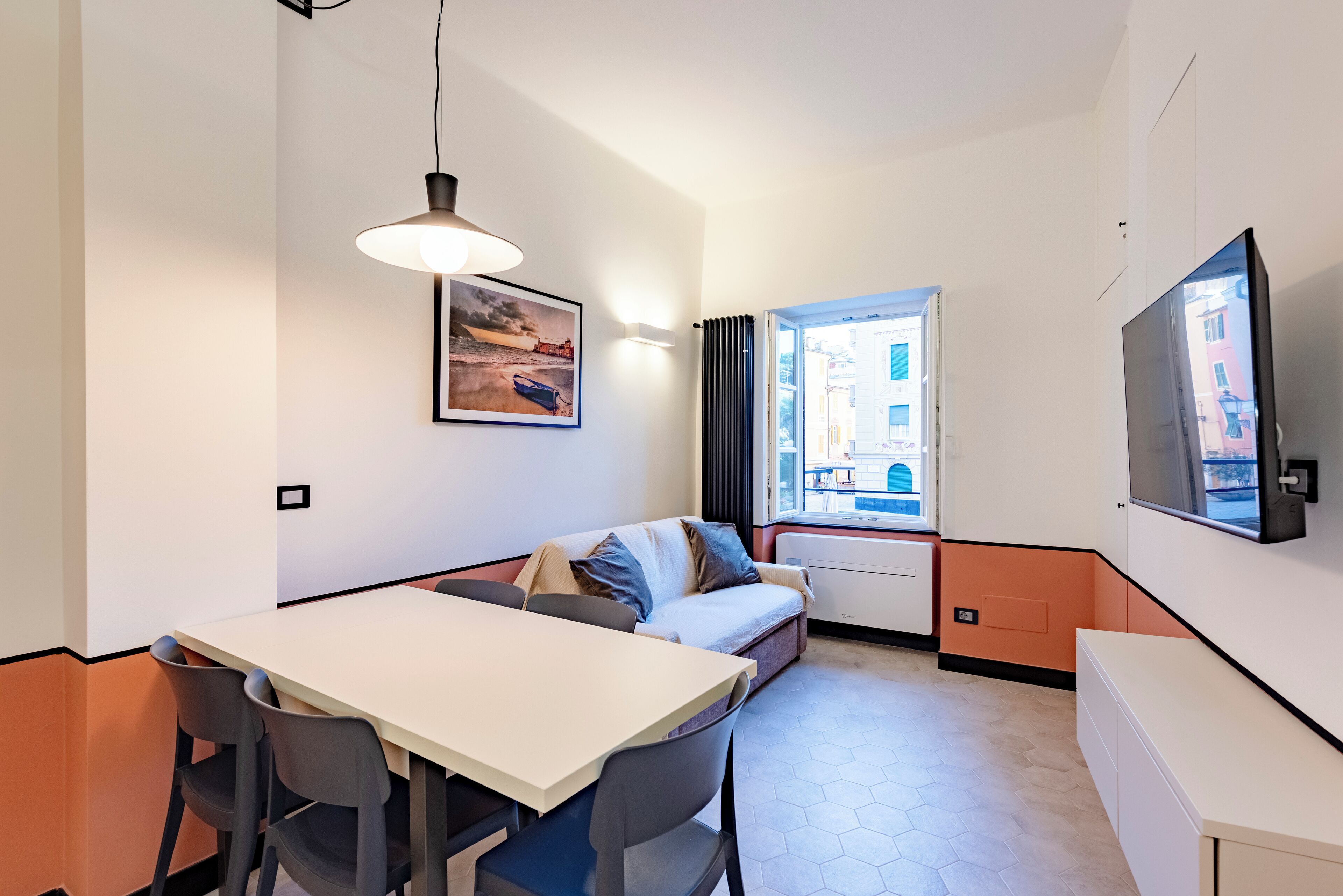 Un espace de vie compact comprenant une table à manger, deux chaises, un canapé, un téléviseur et une fenêtre donnant sur des immeubles.
