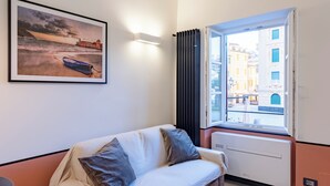 A room with a white sofa, a framed picture of a coastal scene, a window with a view of buildings, and a radiator.