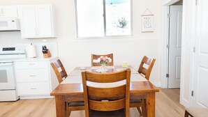Un coin repas avec une table et des chaises en bois, une cuisine blanche dotée de placards et d’appareils électroménagers, et une fenêtre donnant sur un paysage enneigé.
