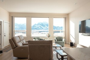 A modern living room with a sofa, chairs, a coffee table, and a TV. Large windows offer a view of mountains.
