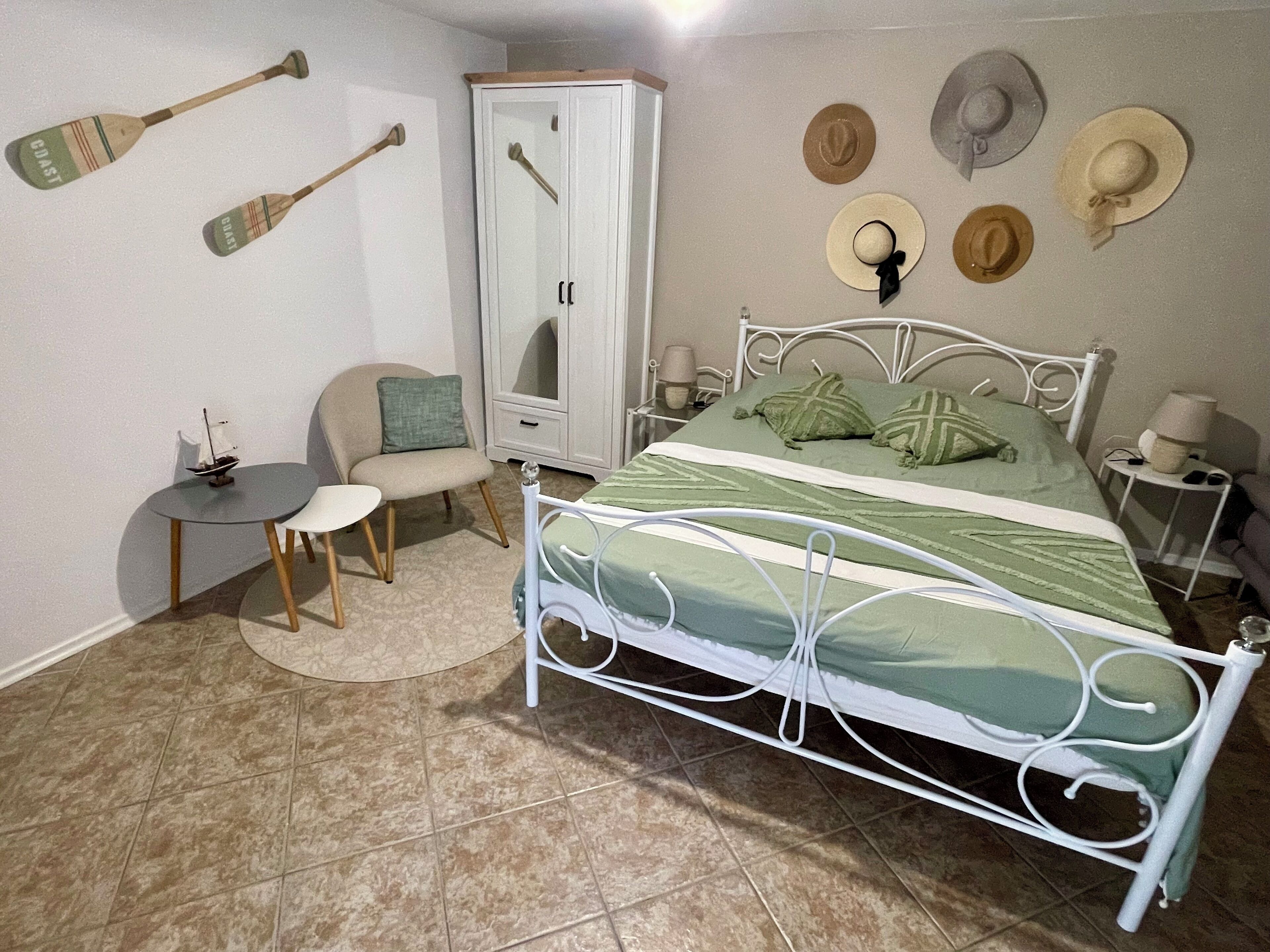 A bedroom with a bed, a chair, a small table, a wardrobe, and several hats on the wall.