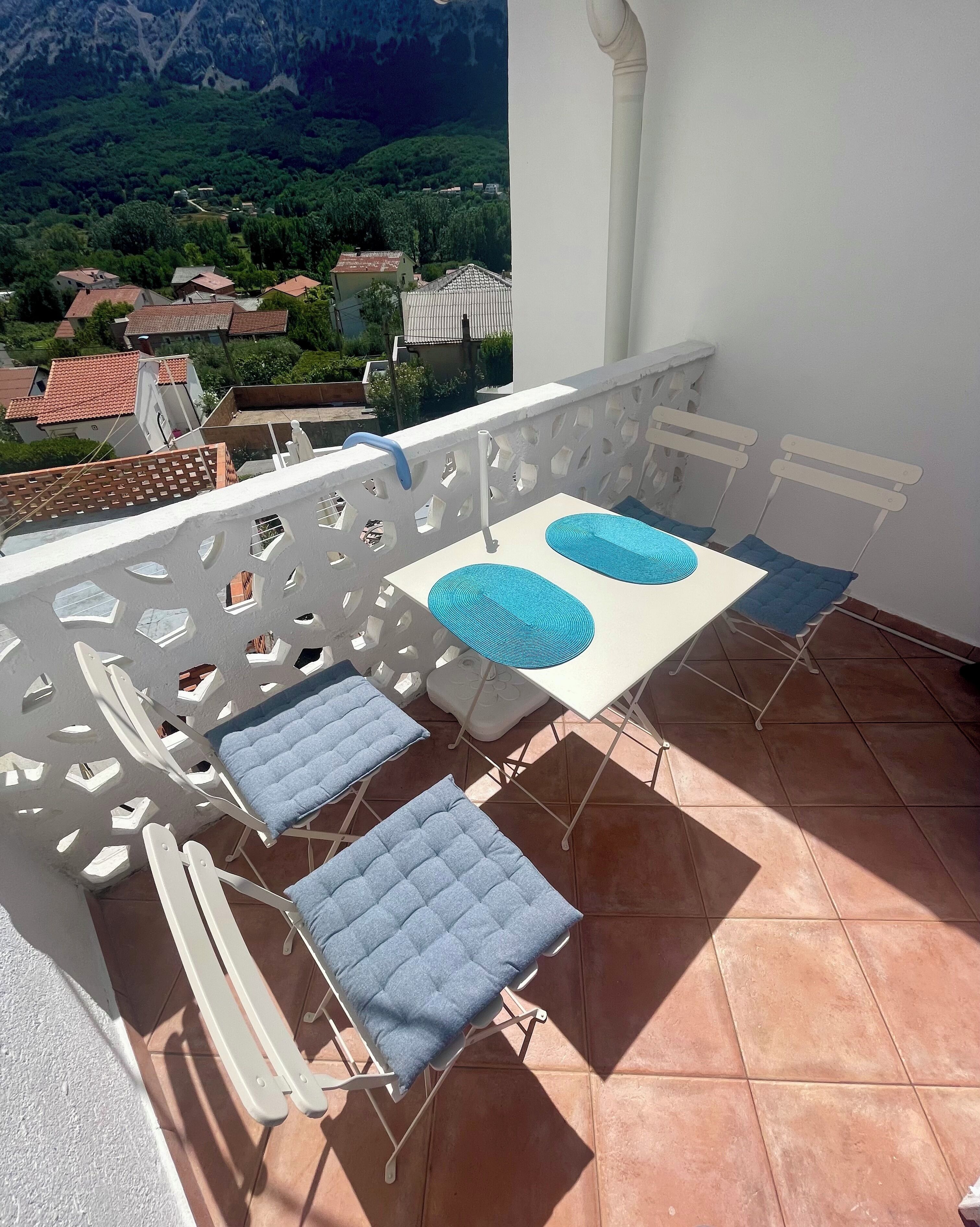 Un balcon avec une table blanche et quatre chaises bleues dotées de coussins, offrant une vue sur un paysage montagneux.