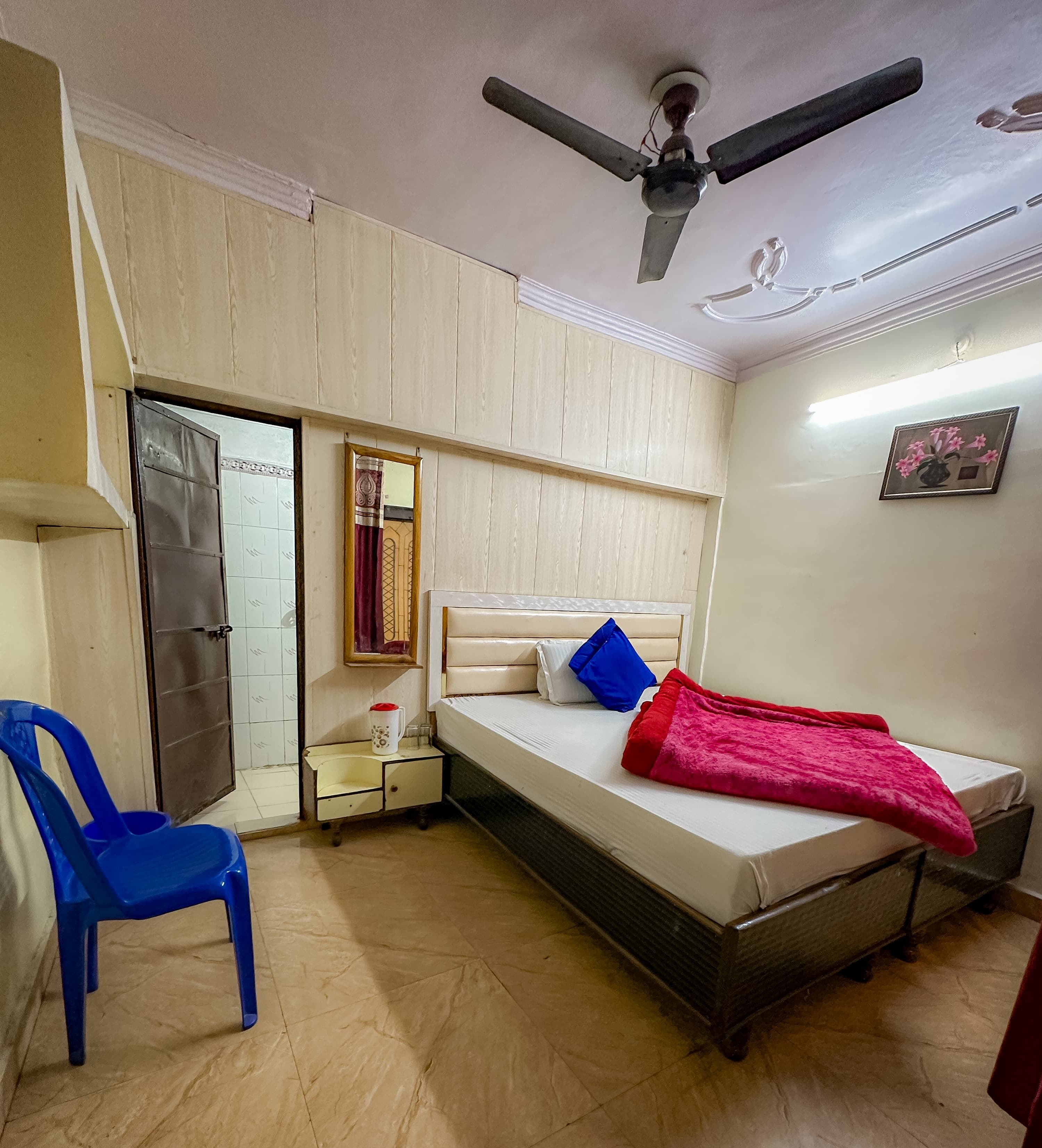 Ein Schlafzimmer mit einem Bett, einem Deckenventilator, einem Nachttisch, einem blauen Stuhl, einer Tür und einem an der Wand hängenden Bild.