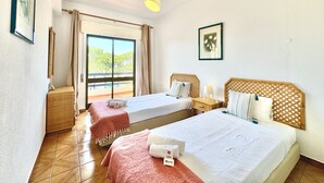 Habitación de hotel con dos camas, un ventanal con vistas a árboles, una lámpara en el techo y un cuadro en la pared.