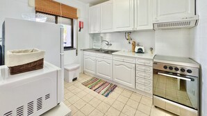 Cocina compacta con muebles blancos, un refrigerador, una estufa y un ventanal con persianas.