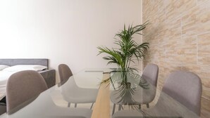 Ein modernes Hotelzimmer mit einem Glastisch, zwei grauen Stühlen, einem Holznachttisch, einer Topfpflanze und einem Bett mit weißen Bettwäsche.