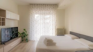 Ein Hotelzimmer mit Bett, Fernseher auf einem Schrank, Heizkörper und einem Fenster mit leichtem Vorhang.