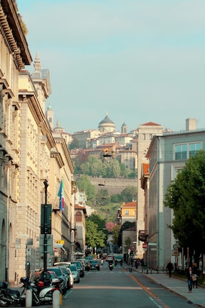 A street lined with historic buildings, a clear view of a hilltop church, and parked cars along the sidewalk.