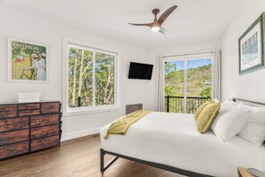 Un dormitorio con cama, cómoda, televisor y ventanas amplias con vistas a árboles.