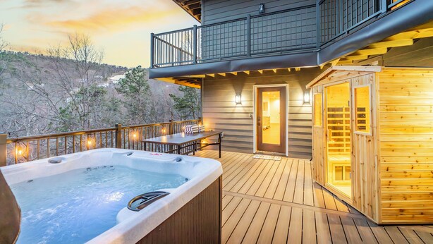Terraza de madera con jacuzzi, sauna y zona de comedor con iluminación exterior.