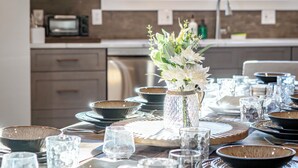 Una cocina luminosa con electrodomésticos modernos, una mesa de comedor dispuesta con varios cuencos y vasos, y un florero con flores en la encimera.