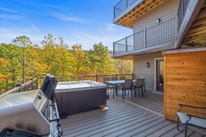 Una terraza de madera con jacuzzi, mobiliario exterior y vistas a árboles otoñales.