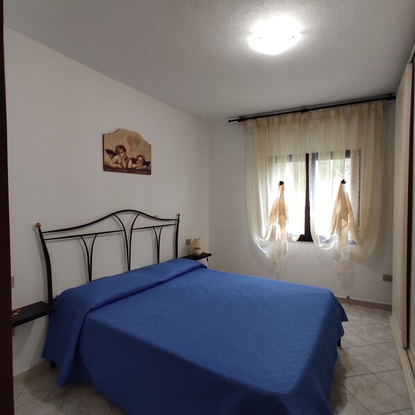 A bedroom with a bed, a nightstand, a lamp, a painting, and a window with curtains.