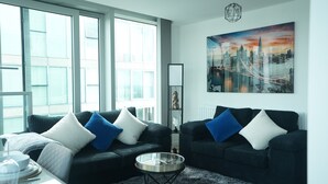 Een moderne woonkamer met twee donkere banken, een glazen salontafel en een stadslandschap schilderij aan de muur.