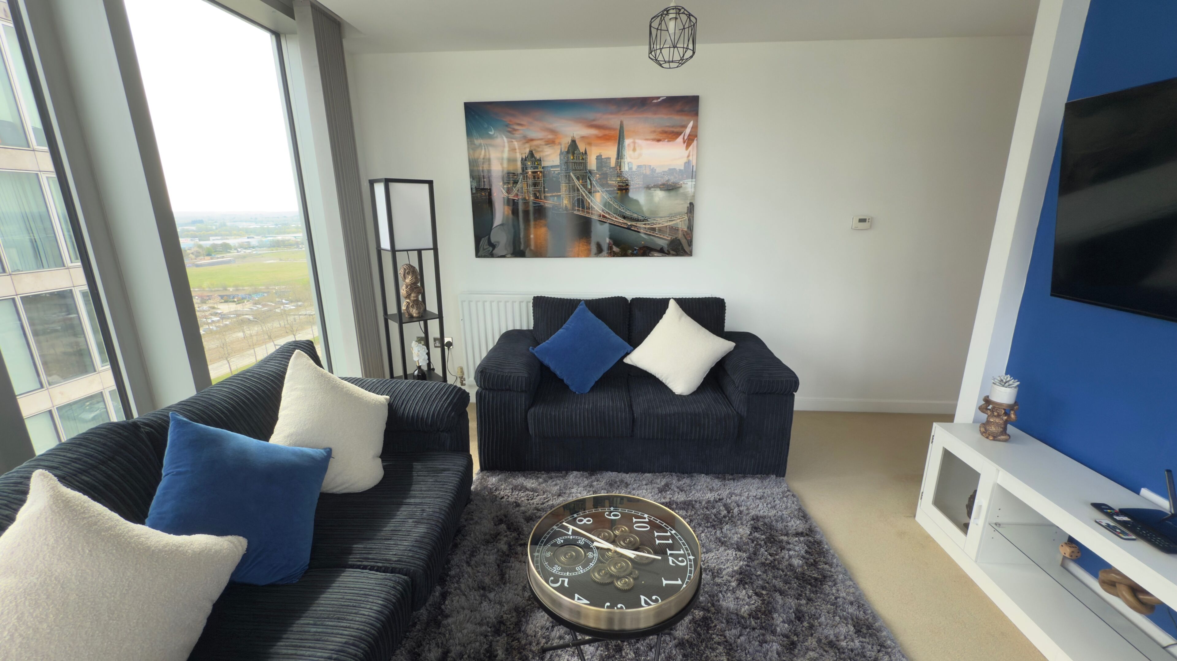 Una sala de estar moderna con un ventanal grande, un sofá negro con cojines azules y blancos, un reloj redondo y un cuadro de un paisaje urbano.