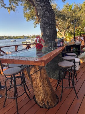 A wooden deck with a bar area featuring stools, a glass-top table, and a large tree trunk as a central support.