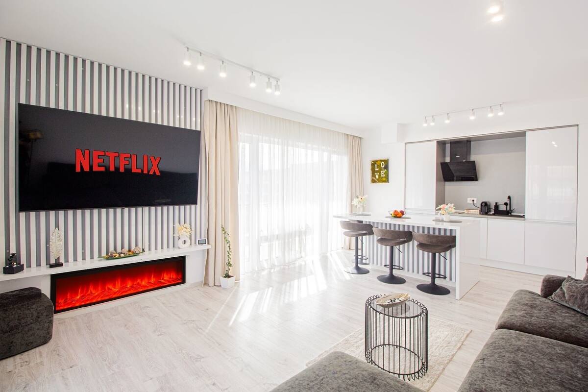 A modern living room with a large flat-screen TV displaying "NETFLIX," a kitchen area, and a cozy seating arrangement.