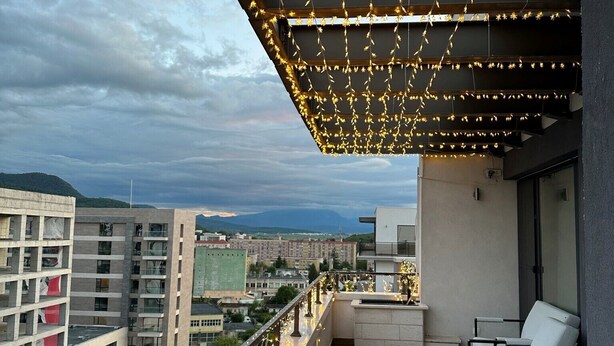 Een balkon met een pergola, versierd met lichtjes, met uitzicht op een stadsgezicht met gebouwen en een bergketen in de verte.