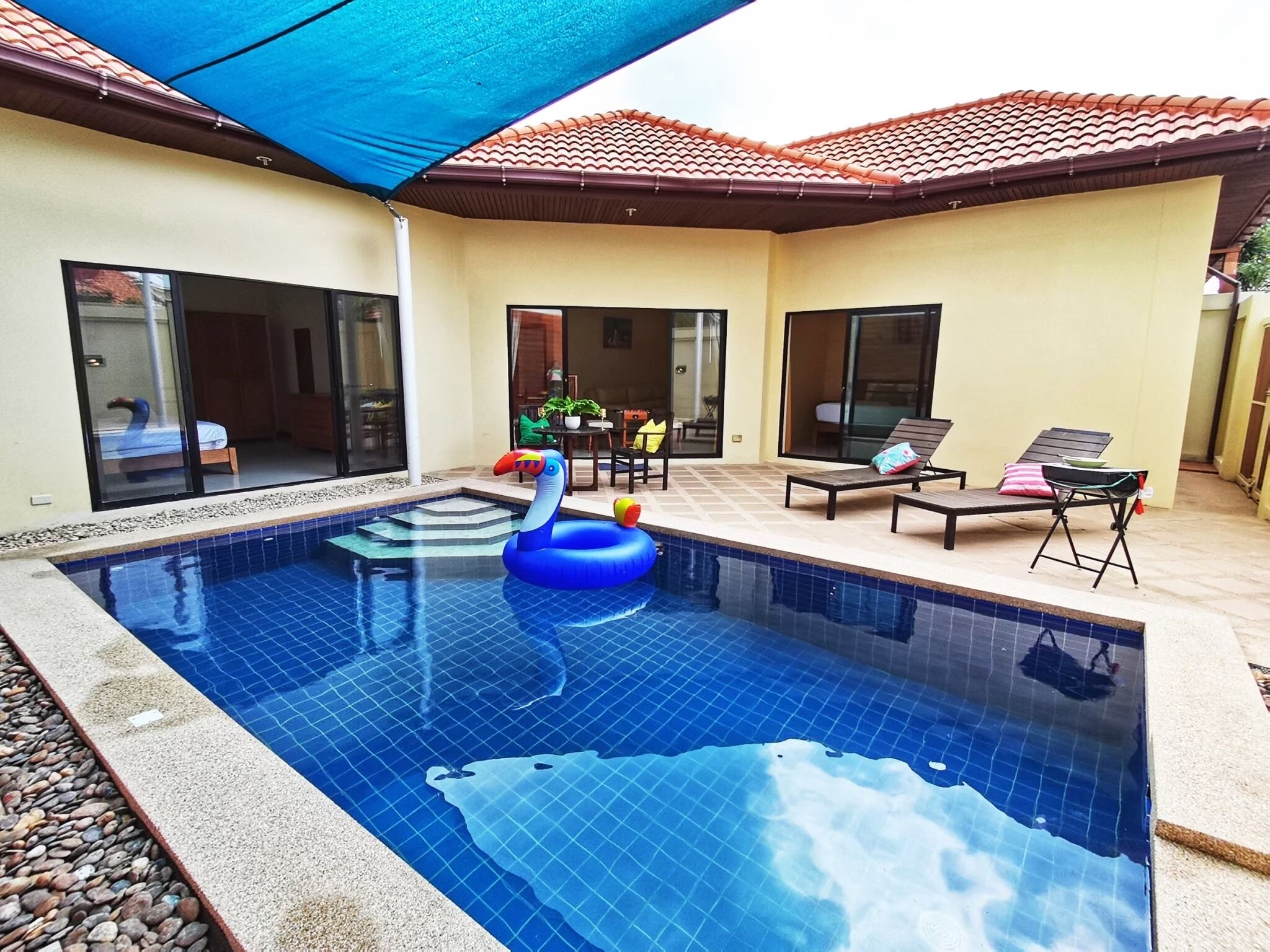 Une piscine avec un flamant rose gonflable bleu, entourée d’un patio aménagé avec des meubles d’extérieur et un espace salon couvert.