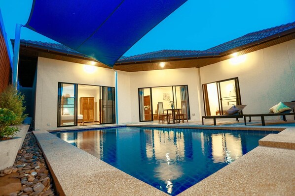 Ein moderner Poolbereich im Freien mit einem klaren, blauen Pool, einer überdachten Sitzgruppe und einer Terrasse mit Tisch und Stühlen.