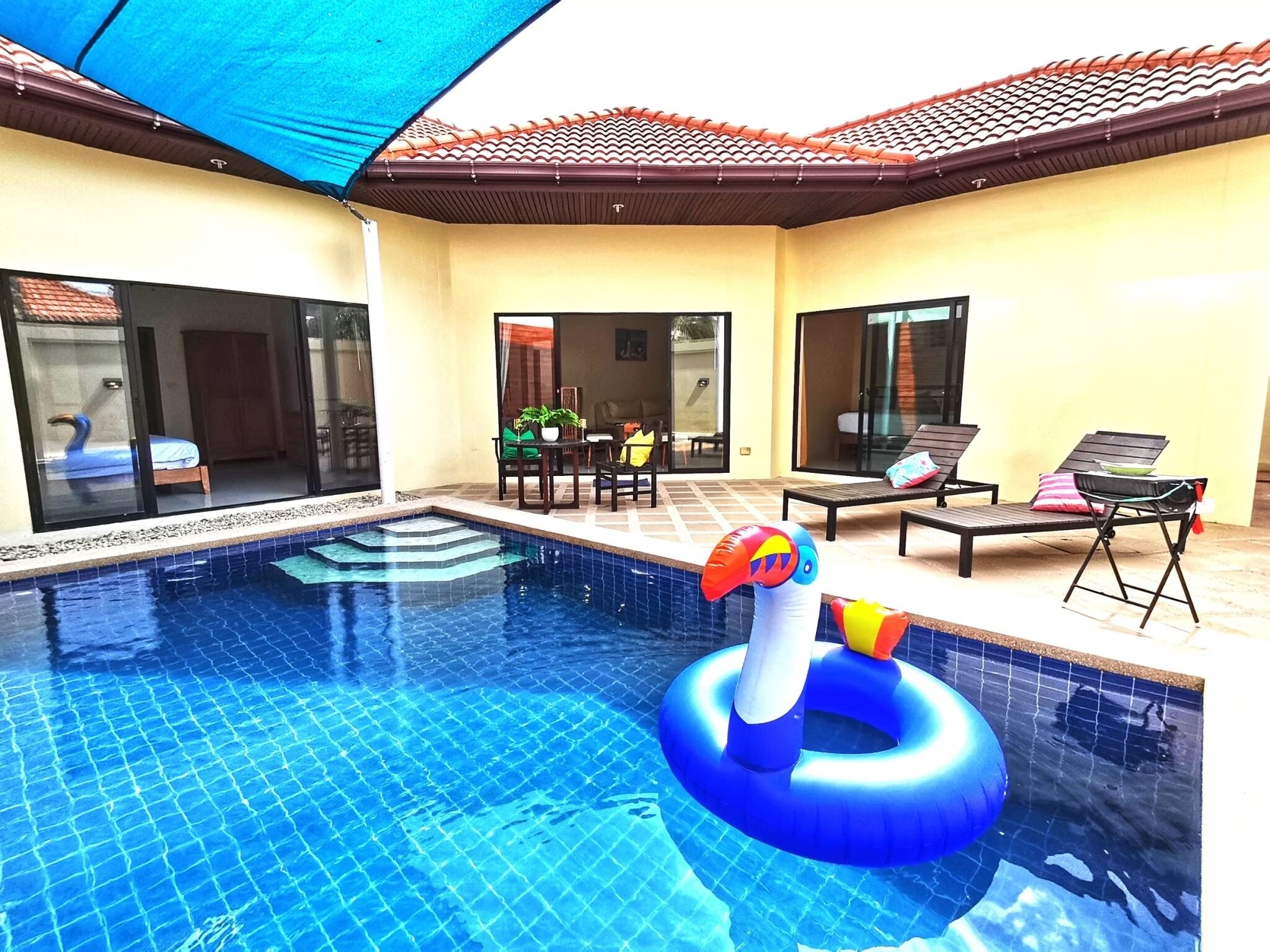 A swimming pool with a blue tiled floor, a lounge area with chairs and a table, and a covered patio with a red-tiled roof.