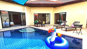 A swimming pool with a blue tiled floor, a lounge area with chairs and a table, and a covered patio with a red-tiled roof.