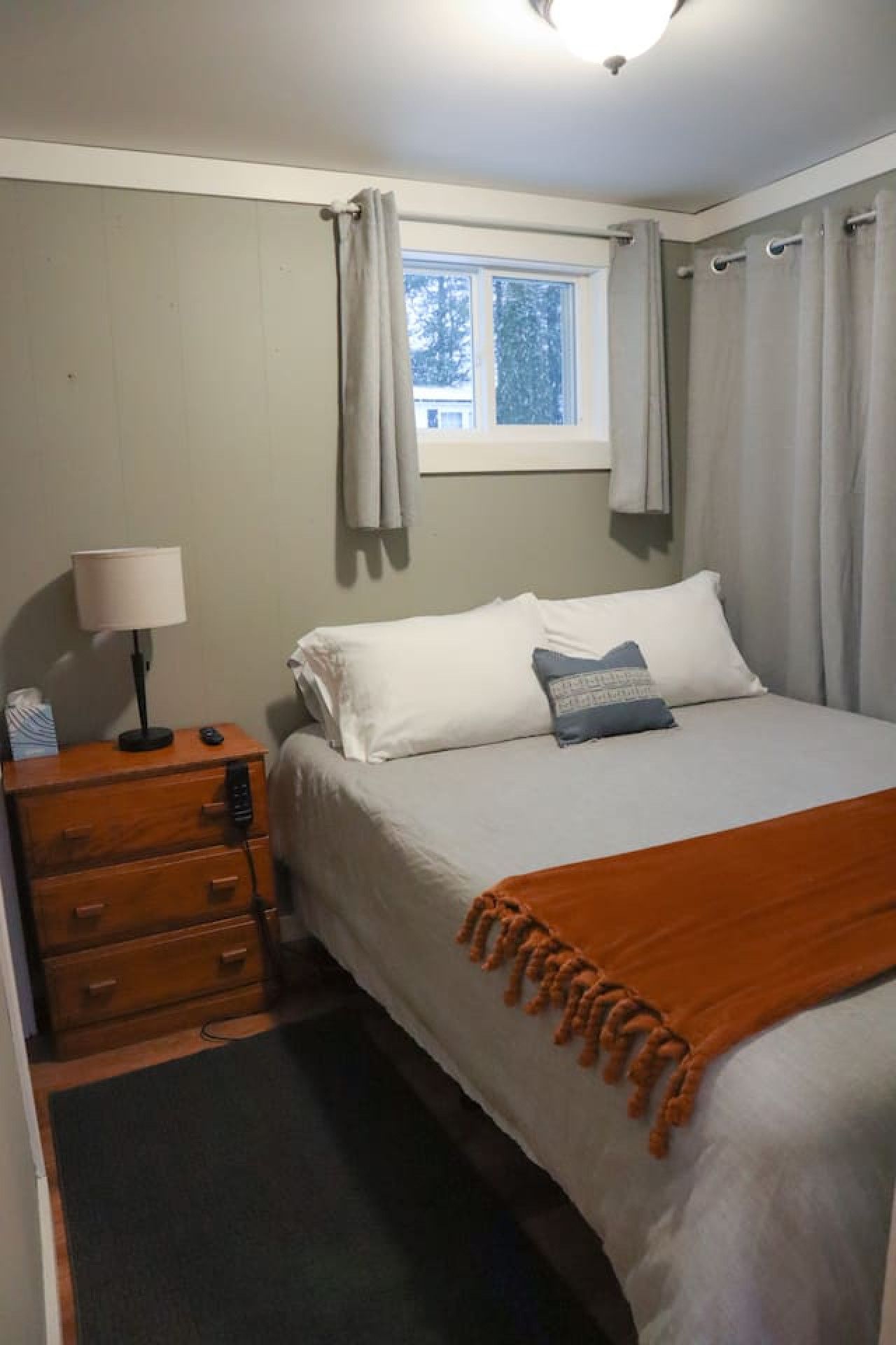 Ein Schlafzimmer mit einem Bett, einem Nachttisch mit Lampe, einem Fenster mit Vorhängen und Blick auf Bäume.