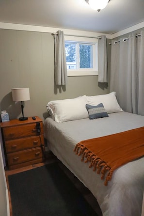 Ein Schlafzimmer mit einem Bett, einem Nachttisch mit Lampe, einem Fenster mit Vorhängen und Blick auf Bäume.