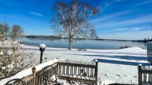 A snow-covered landscape with a bench, a tree, and a body of water under a clear sky. - Modern Lakeside Home (Glenburn)