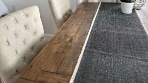 Un comedor con mesa de madera, sillas acolchadas, alfombra tejida, una planta colgante y una cesta de alambre con planta.