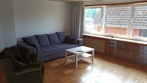 Una sala de estar con un sofá gris, una mesa de centro blanca y un sillón verde. Hay una ventana con vista a un edificio de ladrillo.