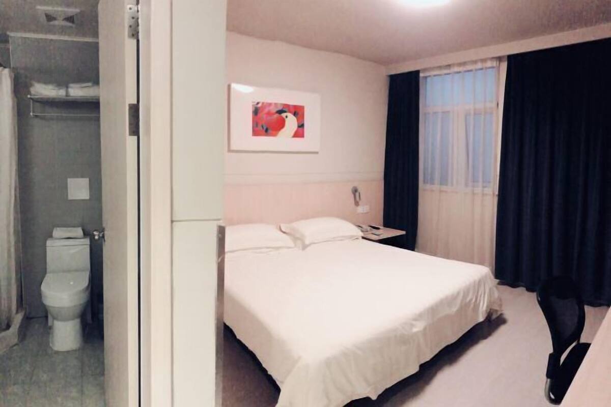 A hotel room with a bed, a bedside table, a window with curtains, a toilet, and a shower area.