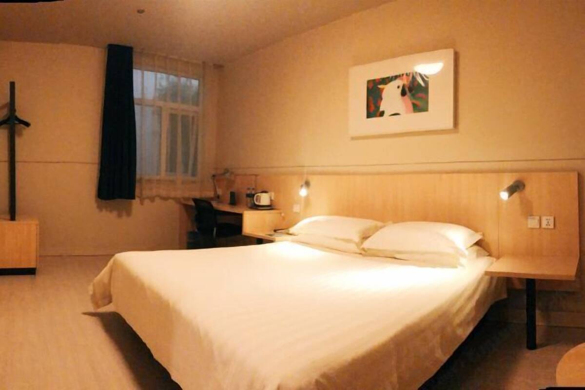 A hotel room with a large bed, a desk with a chair, a window with curtains, and a wall-mounted picture.