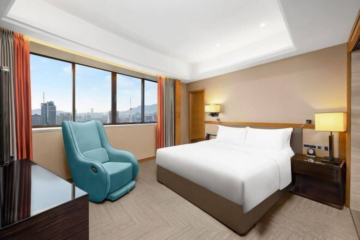 Ein Hotelzimmer mit Bett, Schreibtisch, Stuhl, Nachttisch und Fenster mit Stadtblick.