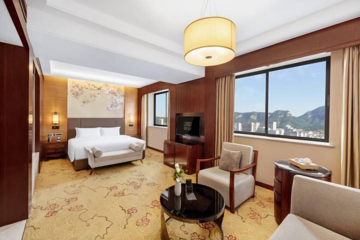 Ein Hotelzimmer mit Bett, Fernseher, Schreibtisch, Stuhl, kleinem Tisch und einem großen Fenster mit Blick auf die Berge.