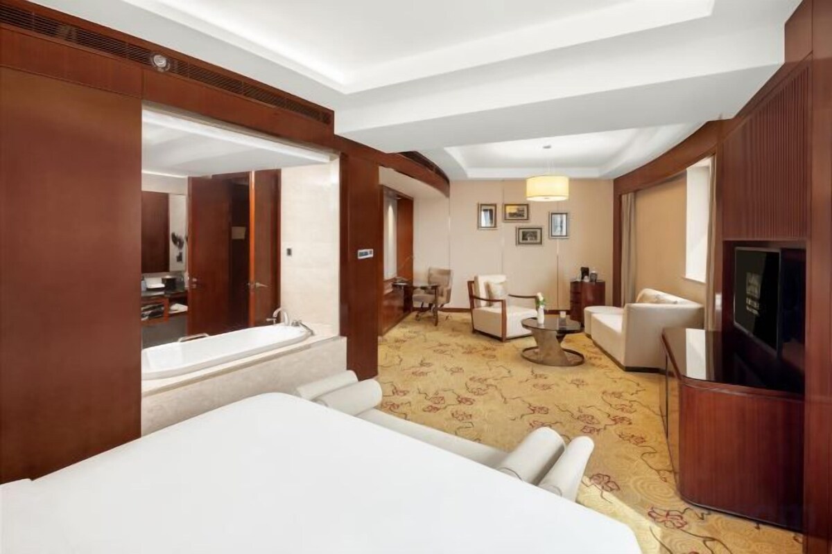 A hotel room with a large bed, a sitting area with a sofa and chairs, a television, and a bathroom with a bathtub.