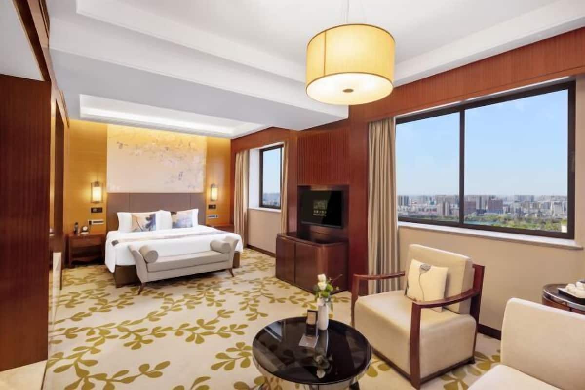 Ein Hotelzimmer mit einem Bett, einem Fernseher, einem Couchtisch und einem Sessel. Ein großes Fenster bietet Blick auf die Stadt.