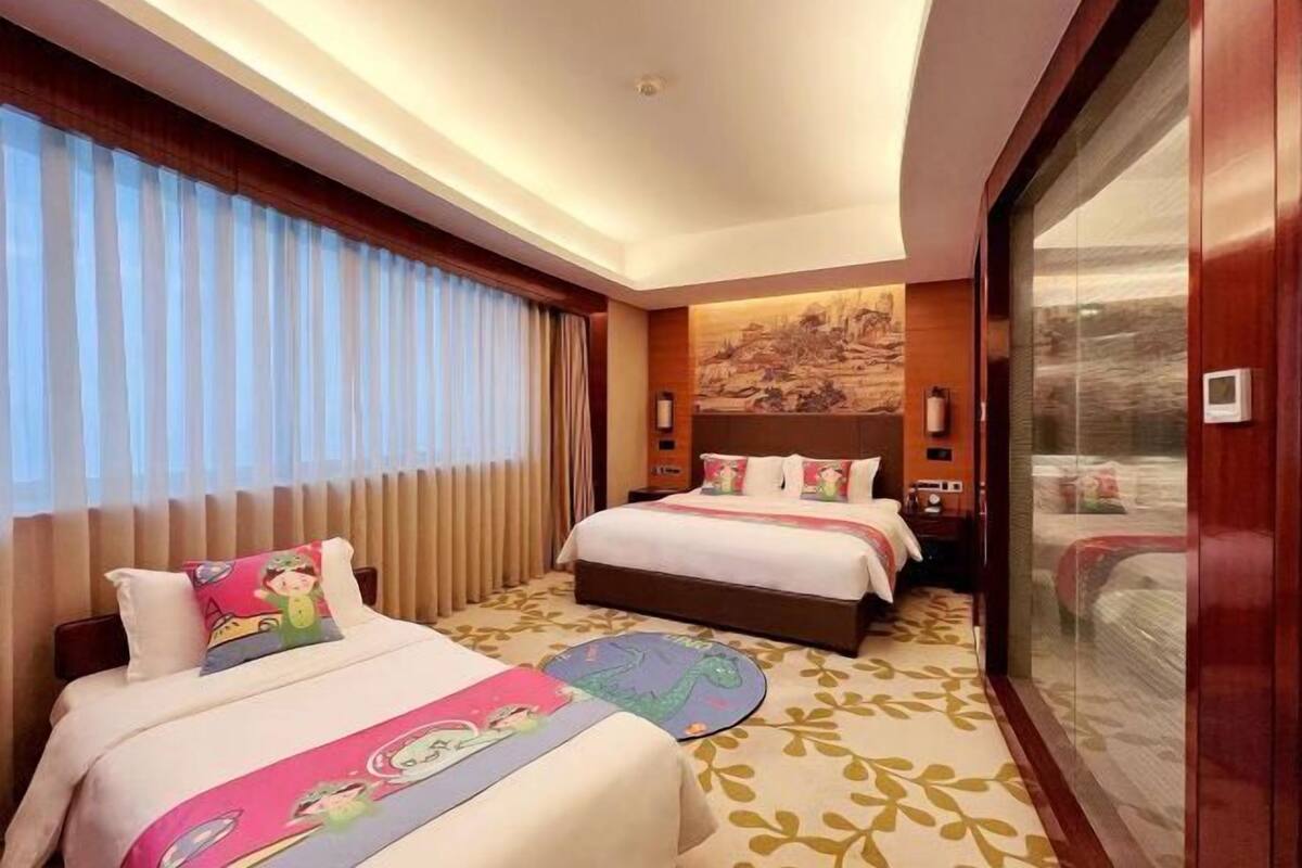 Ein Hotelzimmer mit zwei Betten, einem großen Fenster mit Jalousien, einem Gemälde an der Wand und einer Schiebetür.