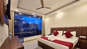 A modern hotel room with a large bed, a flat-screen TV mounted on the wall, a ceiling fan, and a view of a cityscape through a large window. - Aghor Guest House (Dharamshala)