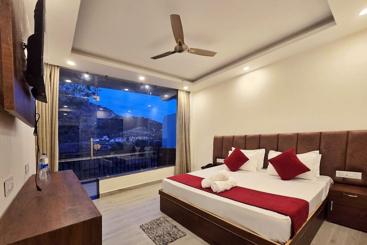 Quarto de hotel moderno com uma cama grande, televisão de tela plana na parede, ventilador de teto e vista para a cidade através de uma janela ampla.