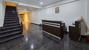 A reception area with a dark wood reception desk, a small stool, a staircase with a handrail, and a ceiling fan. - Aghor Guest House (Dharamshala)